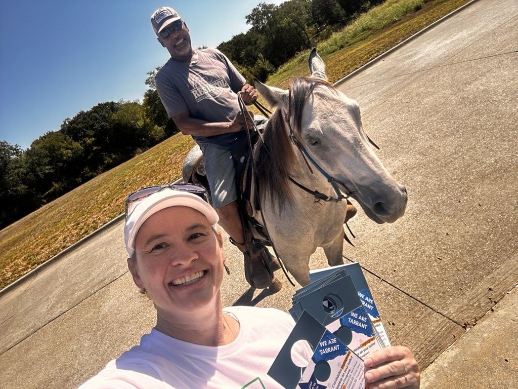 A volunteer in a cap holds campaign literature for a selfie while a man on horseback waits on the road behind her