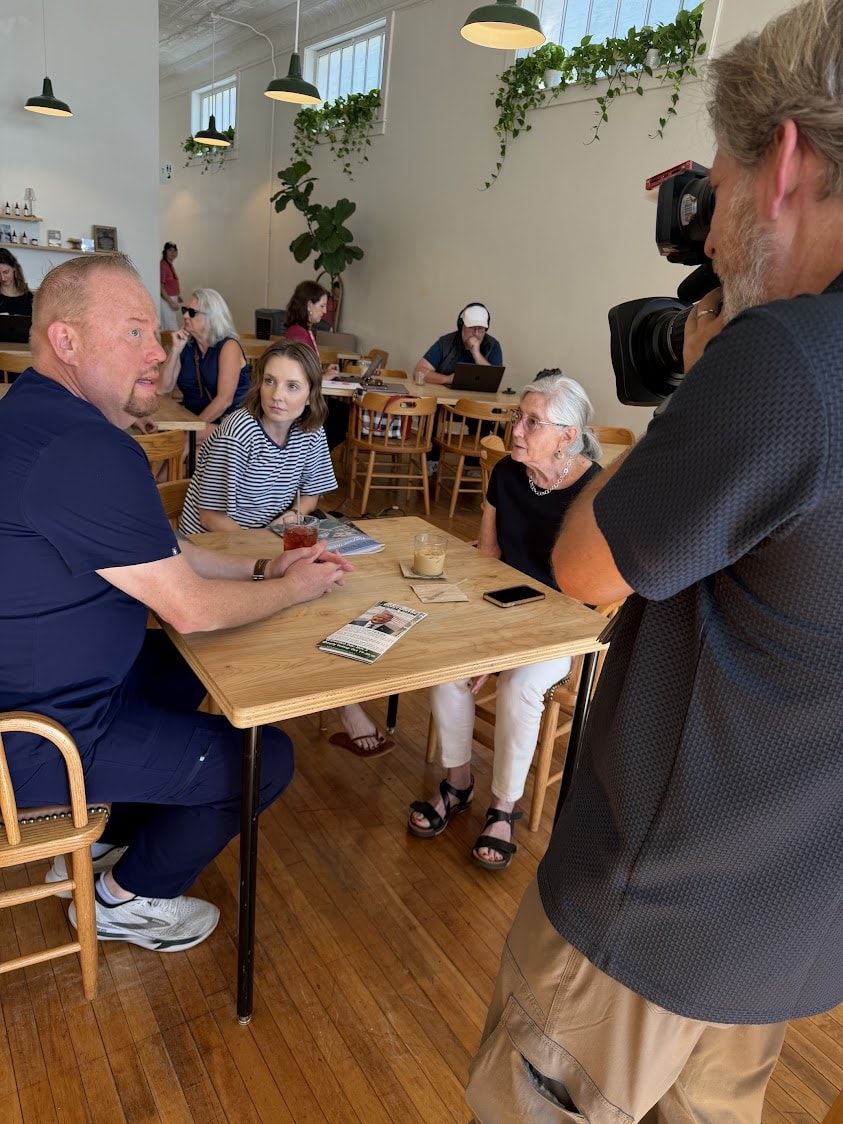 Danny Minton sitting at a cafe table talking with voters while a man films them with a camera